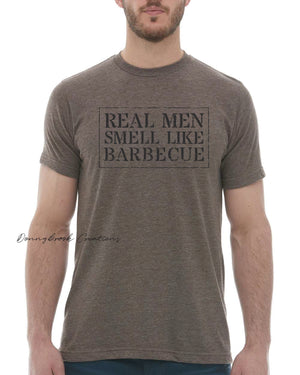 This design features a rustic font that reads "Real Men Smell Like Barbecue" Everyday lightweight smooth & comfy tee.  (Slim Fit). Available in Blue/Green/Grey/Brown